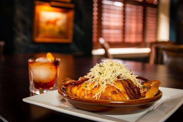 A Hot Brown sandwich in a baking dish on a plate piled high with bacon and cheese and an old fashioned cocktail.